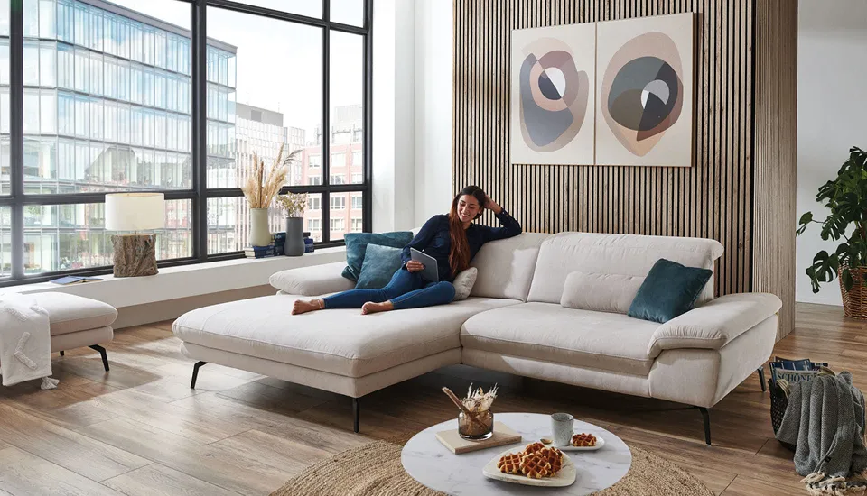 Gemütliches Ecksofa in Beige mit Recamiere und Kissen, platziert in hellem Loft mit großen Fenstern und Holz-Designwand, ideal für entspanntes Wohnen in stilvollem Ambiente.